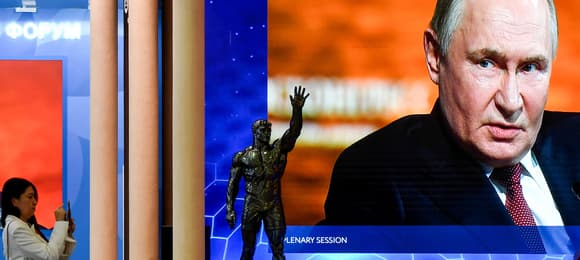 Na wielkim ekranie przemawia mężczyzna, Władimir Putin, na pierwszym planie przechodząca ulicą osoba