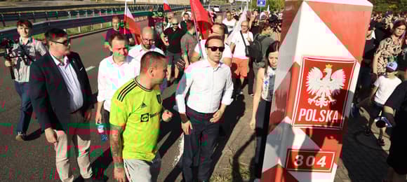 Grupa ludzi na szosie obok słupa granicznego z godłem Polski