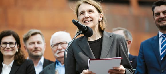 Uśmiechnięta 50-latka z włosami do ramion i w szarej marynarce przed mikrofonem