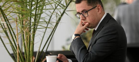 Miłosz Motyka siedzi zamyślony drabie się po podbródku na tle paprotki, trzymając w ręku kubek, zapewne z ciepłym napojem w środku