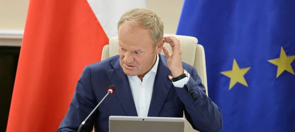 Mężczyzna siedzi, drapie się w głowę, za nim flagi - polska i unijna