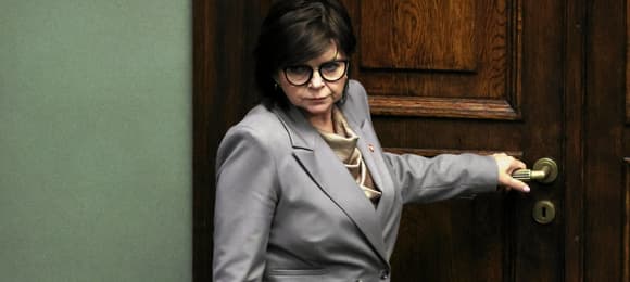 Kobieta z ciemnymi włosami, w okularach, trzyma drzwi za klamkę przed wyjściem.
