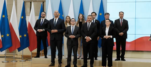 Rząd mężczyzn i kobiet w ciemnych garniturach na tle biało-czerwonych i unijnych flag