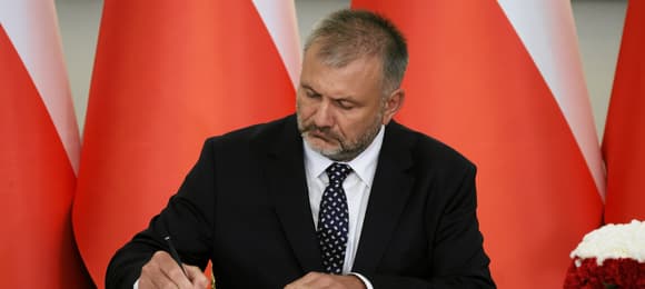 Mężczyzna w garniturze i z zarostem podpisuje dokument. Z tyłu biało-czerwone flagi