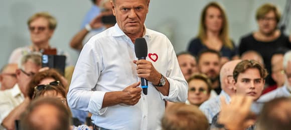 Mężczyzna w białej koszuli z przypinką w kształcie serca mówi do mikrofonu podczas spotkania z ludźmi; w tle widać skupioną publiczność, część osób siedzi, a część stoi