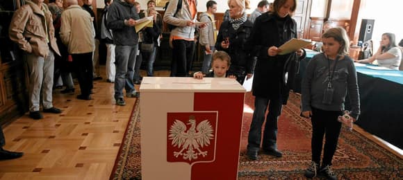 Ludzie w kolejce do biało-czerwonej urny z godłem państwowym