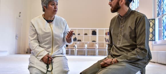 Kobieta z białej tunice i męzczyzna w brązowej tunice i turbanie klęczą na podłodze meczetu i rozmawiają