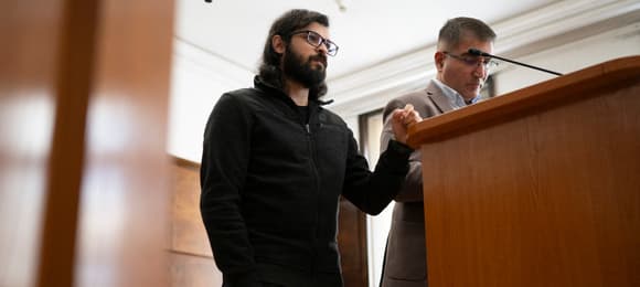 Mężczyzna z długimi czarnymi włosami i brodą, w okularach stoi przy pulpicie dla świadków w sądzie, obok niego drugi mężczyzna.