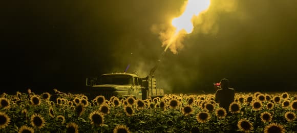 Na nocnym niebie widać rozbłysk pocisku artyleryjskiego wystrzelonego z pojazdu postawionym na polu słoneczników