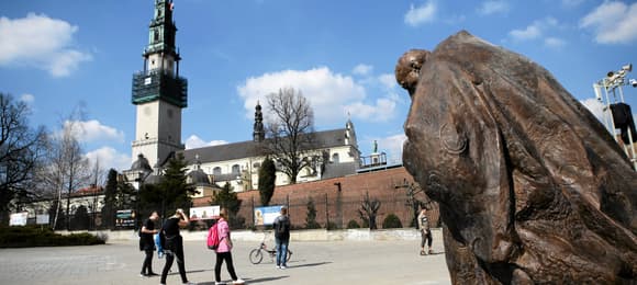 Plac, wysoka kościelna wieża i pomnik klęczącego duchownego.