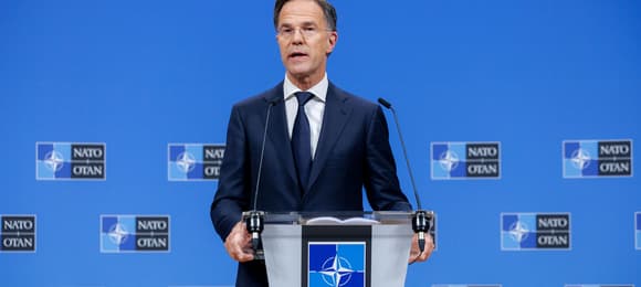 Mężczyzna w garniturze przemawia na konferencji prasowej NATO, stojąc za mównicą z logo Sojuszu na tle niebieskiej ściany z emblematami NATO.