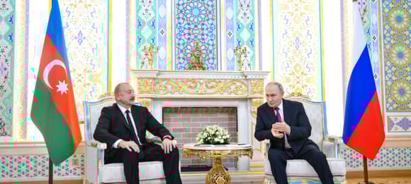 Dwaj starsi mężczyźni w złoconych fotelach. Jeden (Alijew) siedzi rozparty, drugi (Putin) pochula się w jego stronę i tłumaczy