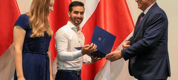 Na zdjęciu jest trzy osoby, po lewej - kobieta, po prawej dwóch mężczyzn. W tle widać flagi biało-czerwone