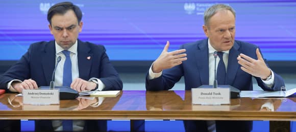 Dwaj mężczyźni w garniturach za stołem, przed każdym tabliczka z nazwiskiem z t