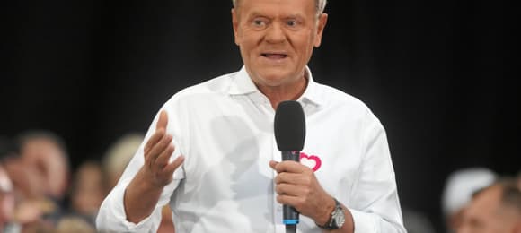 Mężczyzna z blond włosami, w białej koszuli z czerwonym serduszkiem na klatce piersiowej, trzyma mikrofon i przemawia do ludzi.