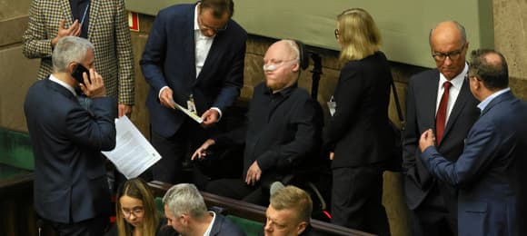 Ludzie w ławach rządowych. Po środku młody mężczyzna na wózku i z aparatem wspomagającym odychanie