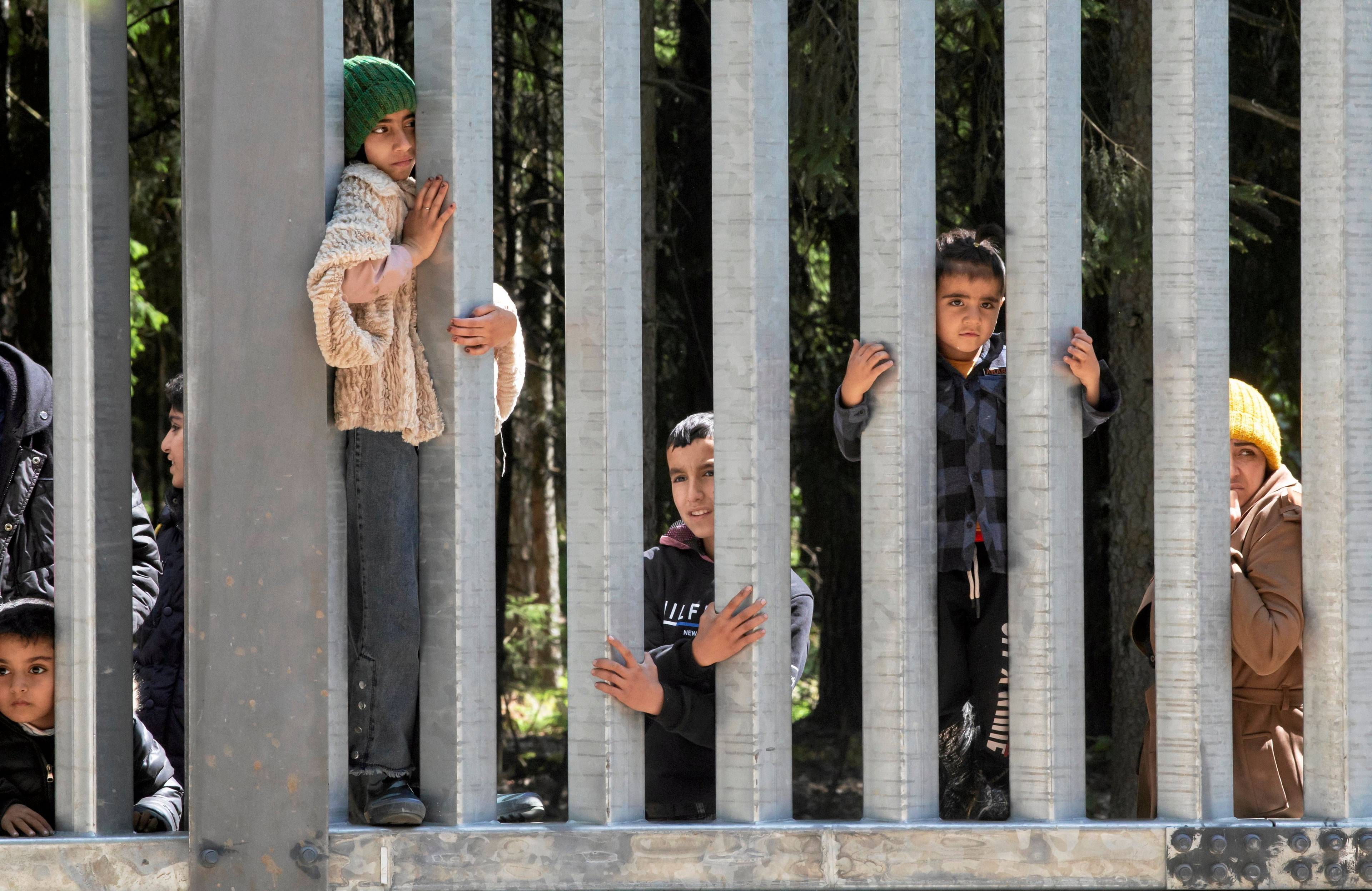 Foto zapory na granicy, między jej prętami widać grupę osób, dorosłych i dzieci