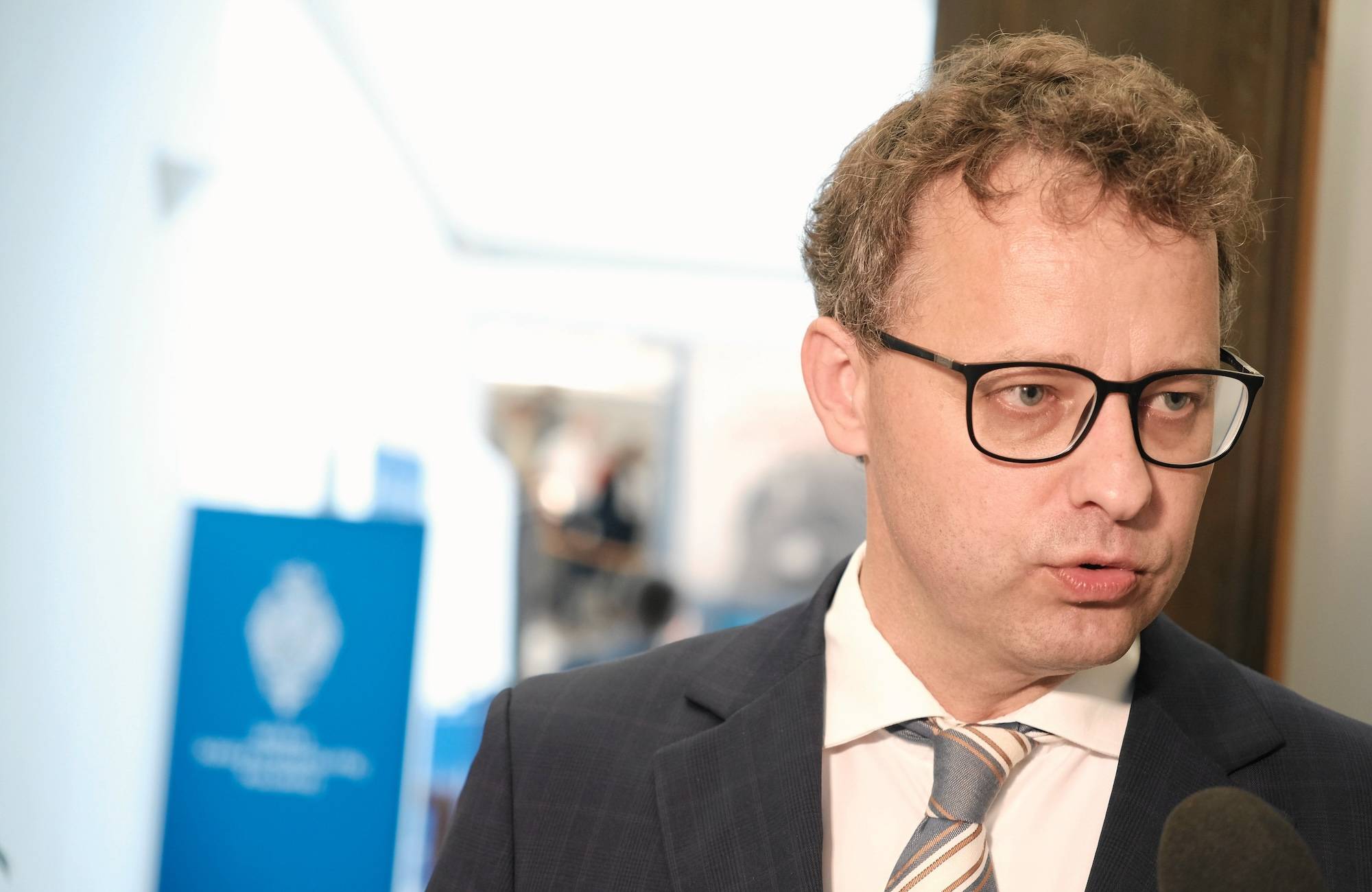 Młody mężczyzna w ciemnej marynarce i krawacie. Nosi okulary w ciemnych, prostokątnych oprawkach