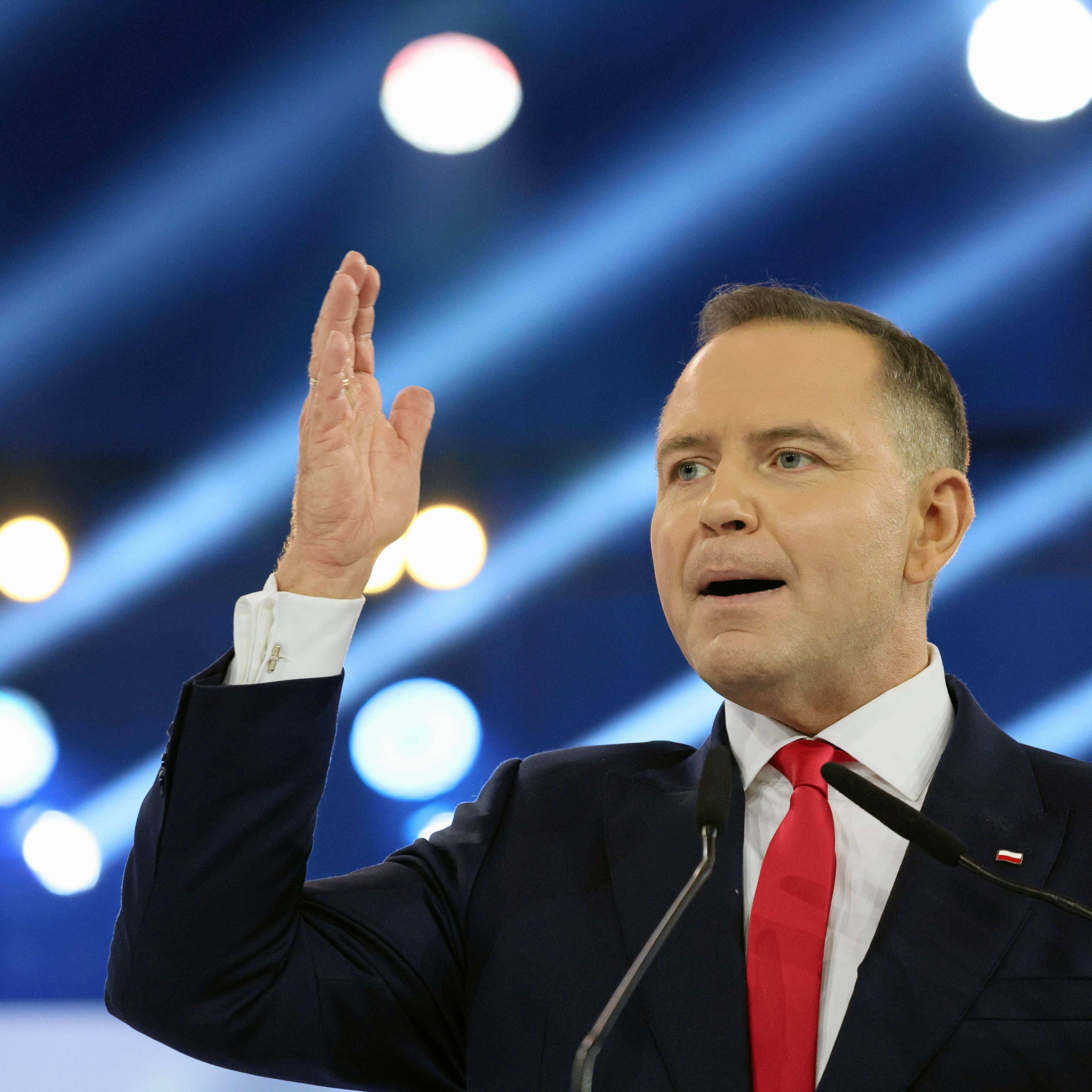 Mężczyzna z uniesioną dłonią, w garniturze i z czerwonym krawatem. Karol Nawrocki, konwencja programowa