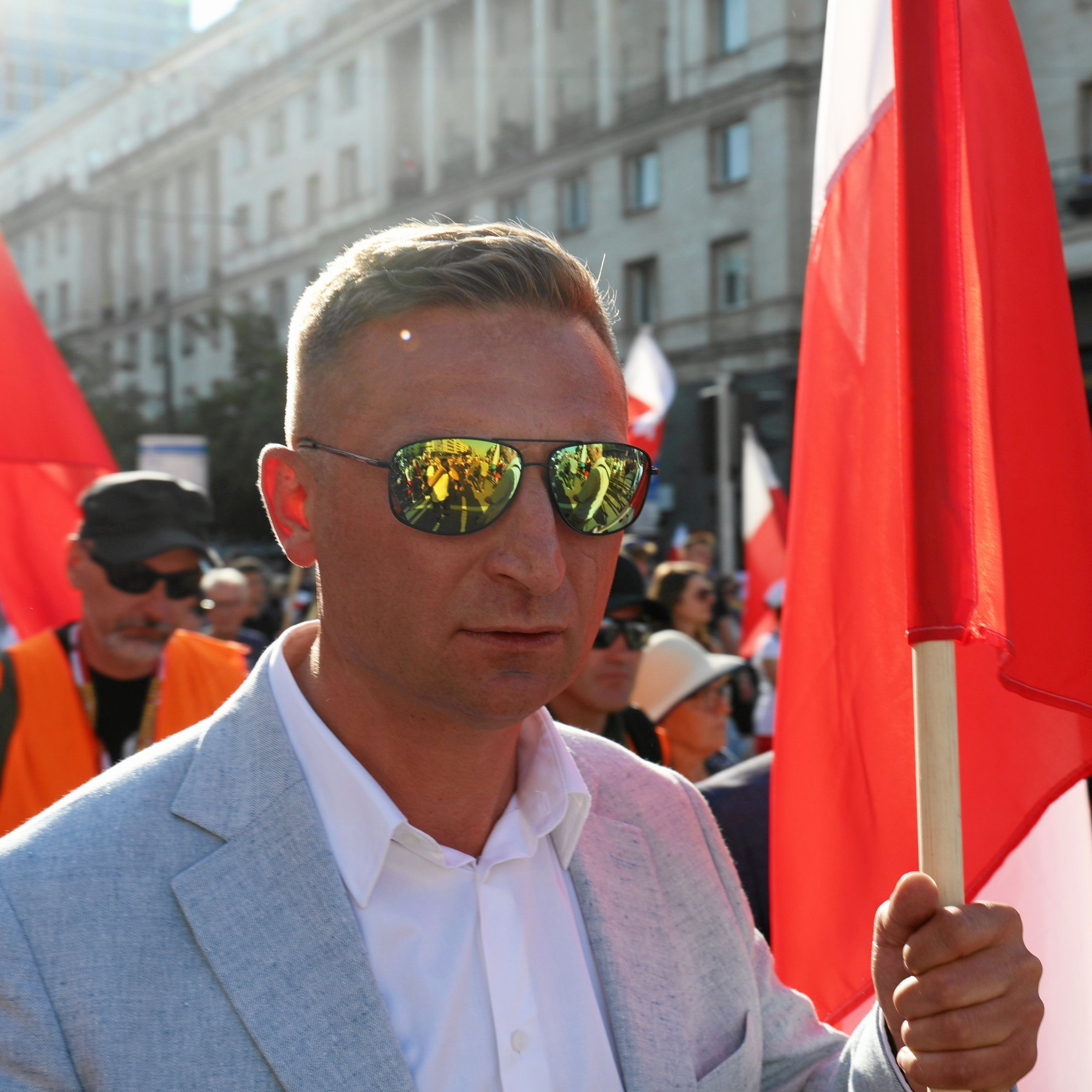 Krótko ostrzyżony mężczyzna w okularach przeciwsłonecznych i błękitnej marynarce maszeruje w tłumie ludzi z biało-czerwoną flagą w ręce