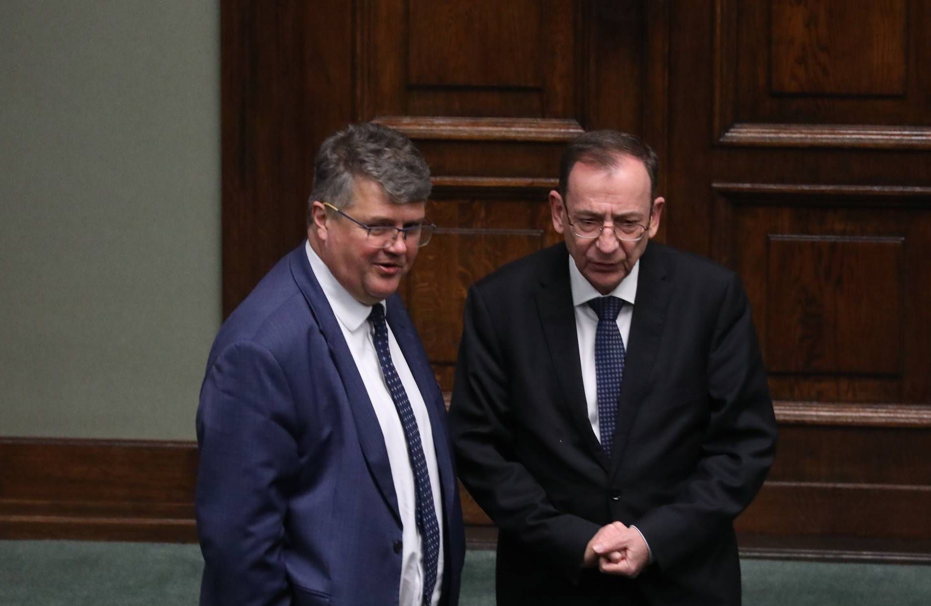 Dwóch mężczyzn w ciemnych garniturach z krawatami obok siebie, w tle ozdobne, masywne, drzwi z ciemnego drewna
