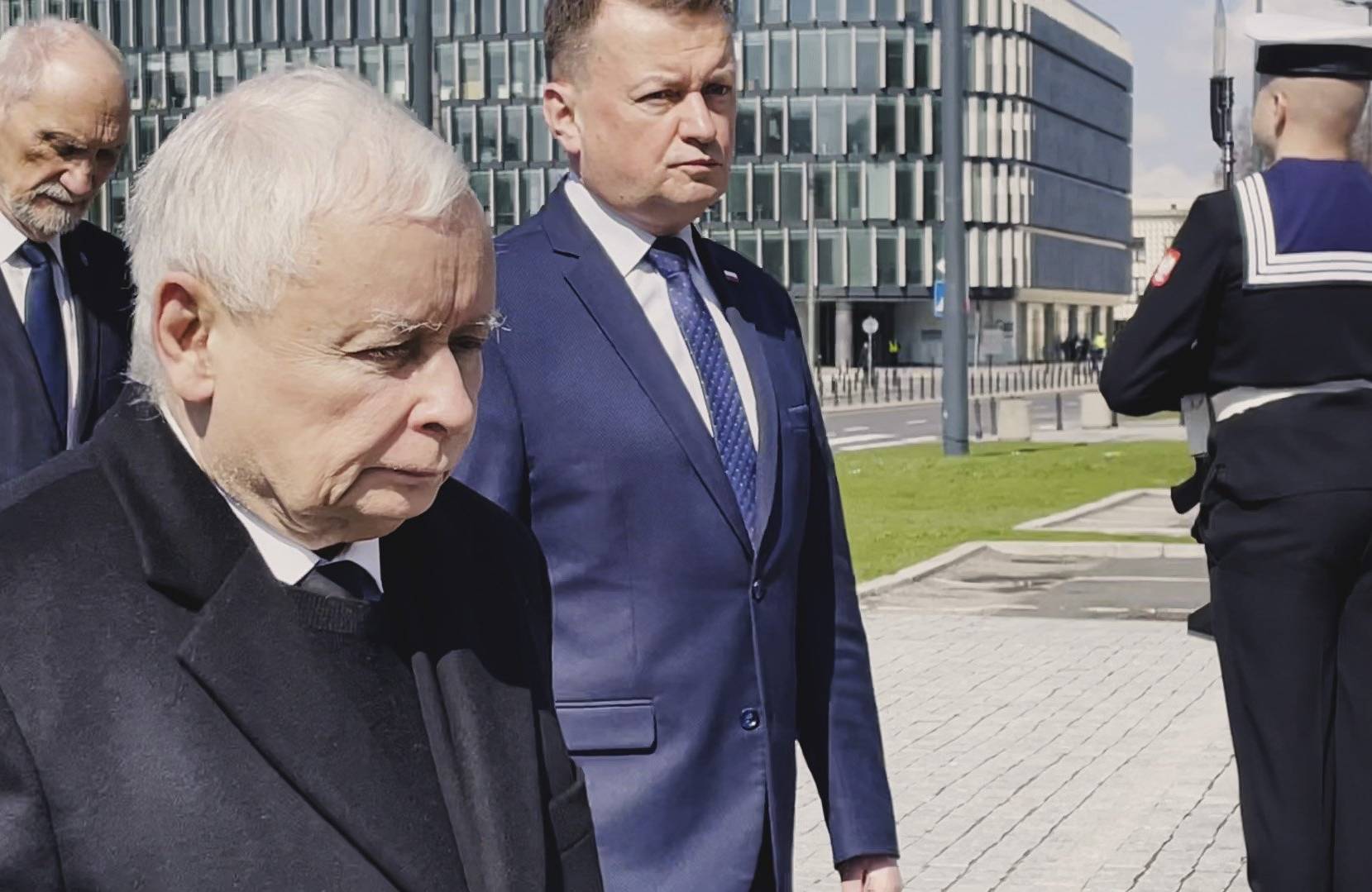 Trzech mężczyzn z ponurymi minami, obok salutujący żołnierz-marynarz