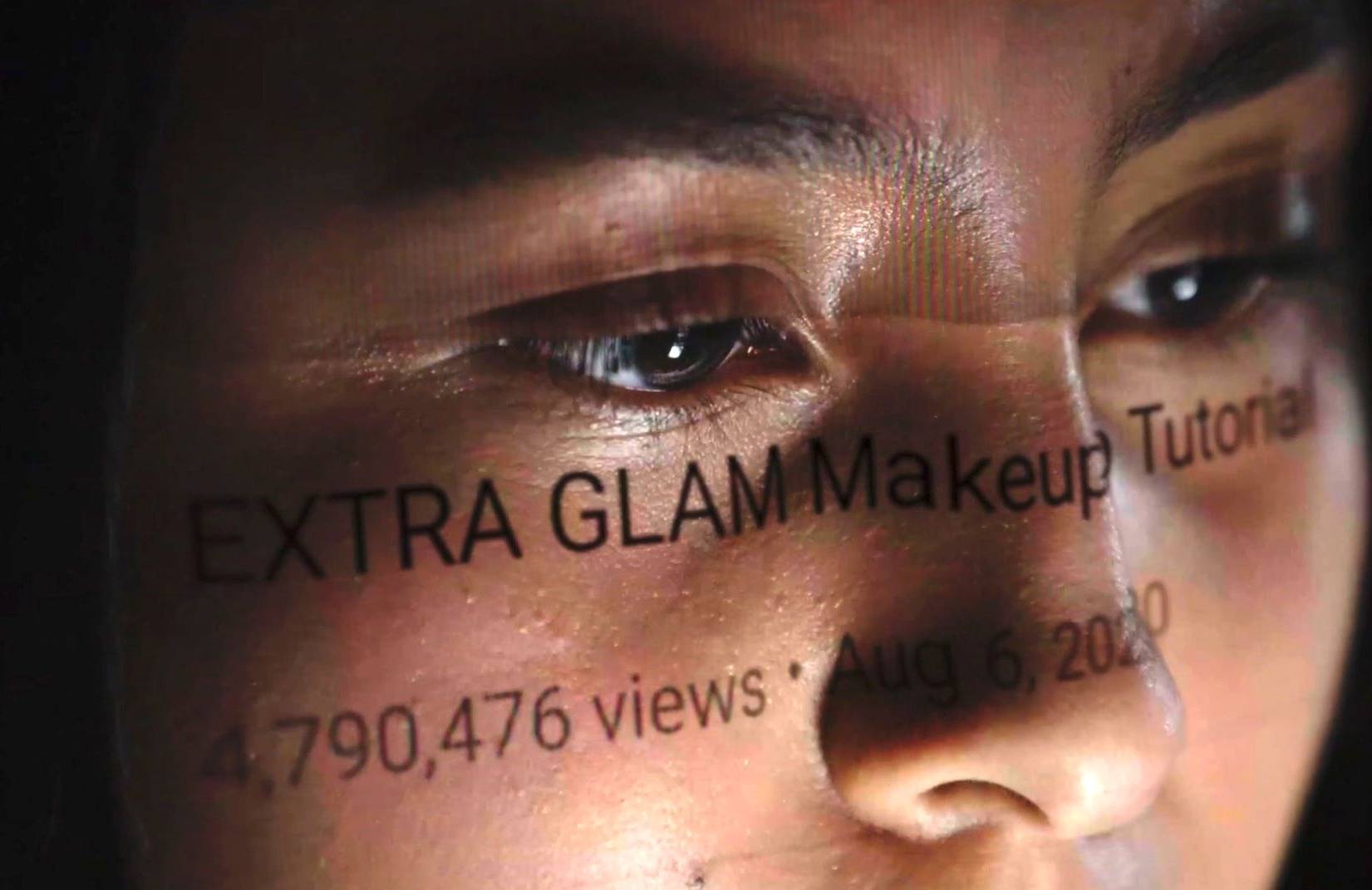 Twarz kobiety, na której odbija się obraz z ekranu komputera z napisem w j. angielskim Extra Glam Make Up Tutorial