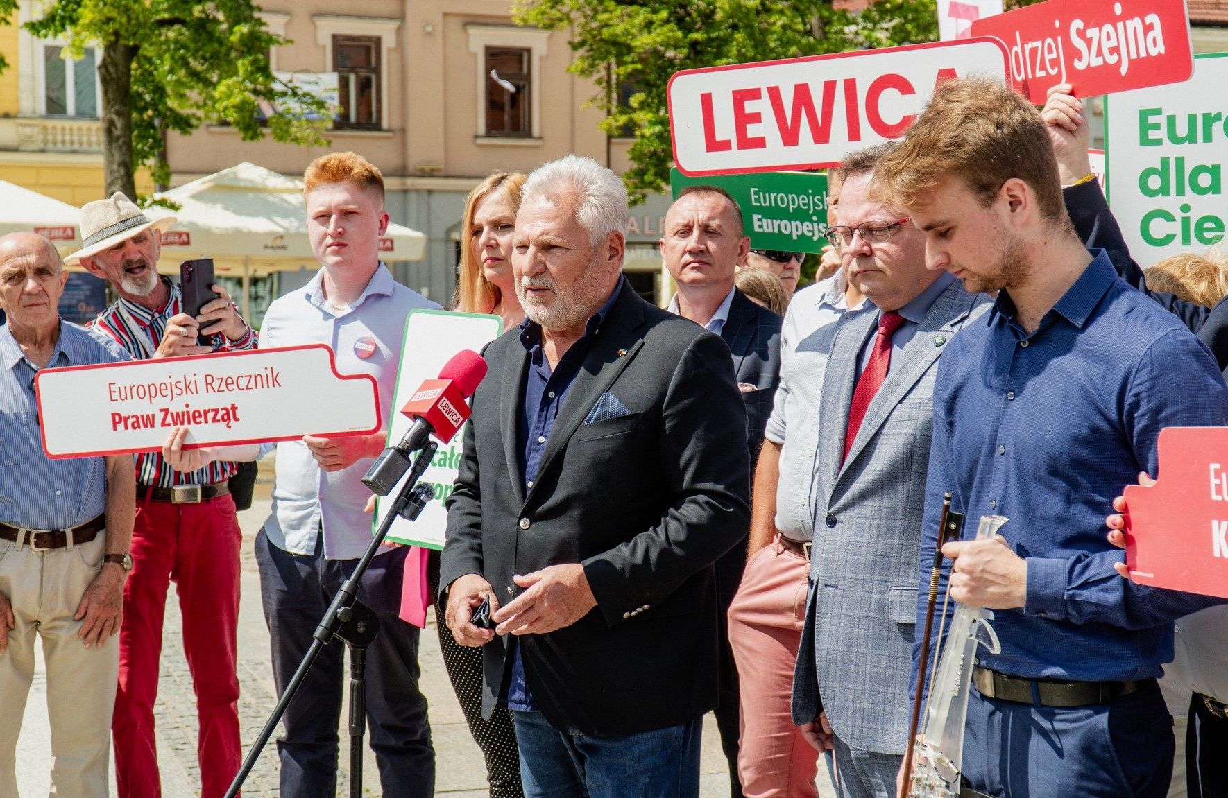 Starszy mężczyzna w siwych włosach, czarnej marynarce, ciemnoniebieskiej koszuli i jeansach przemawia do mikrofonu w otwartej przestrzeni, na placu pod kamienicami w mieści; w tle stoją działacze partii Lewica