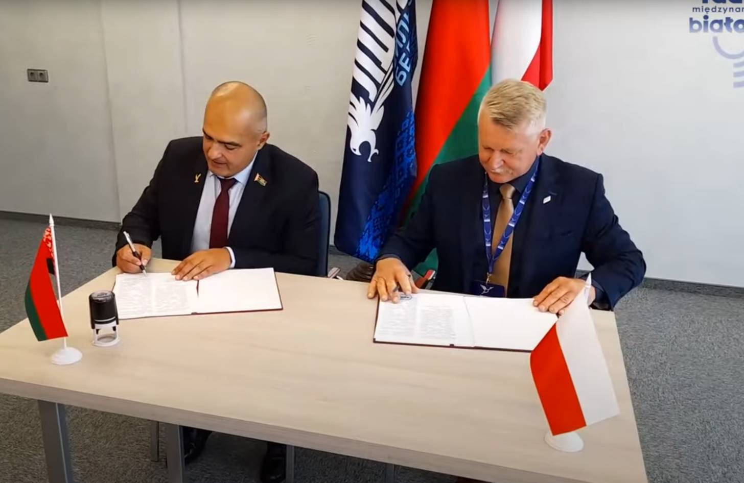 Z prawej Oleg Hajdukiewicz, z lewej - Krzysztof Tołwiński. Dwóch mężczyzn w zaawansowanym wieku średnim, obaj w marynarkach, mężczyzna z lewej łysy, mężczyzna z prawej siwy. Siedzą przy stoliku. Podpisują leżące przed nimi dokumenty. W tle flaga białoruska.
