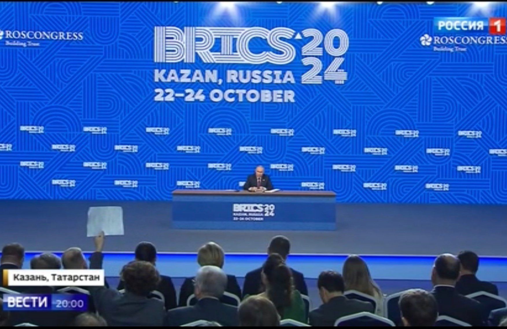 Maleńka sylwetka Putina na tle wielkiego stołu i niebieskiego ekranu z napisem BRICS