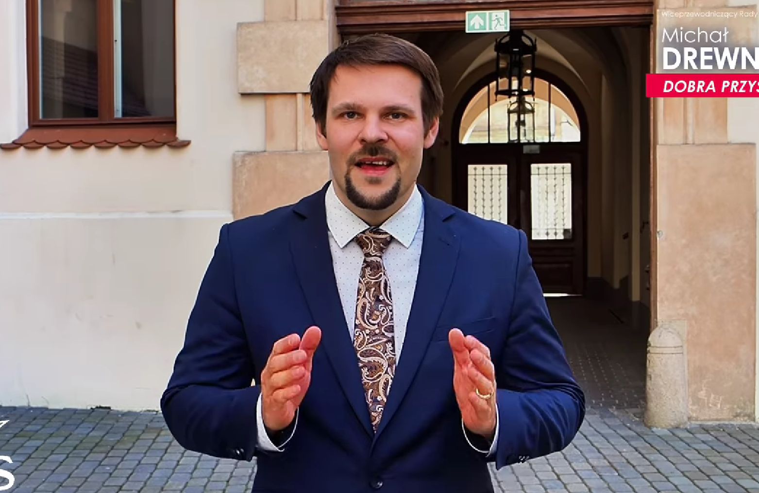 Mężczyzna w garniturze w średnim wieku, z wąsami i brodą, na tle zabytkowej kamienicy