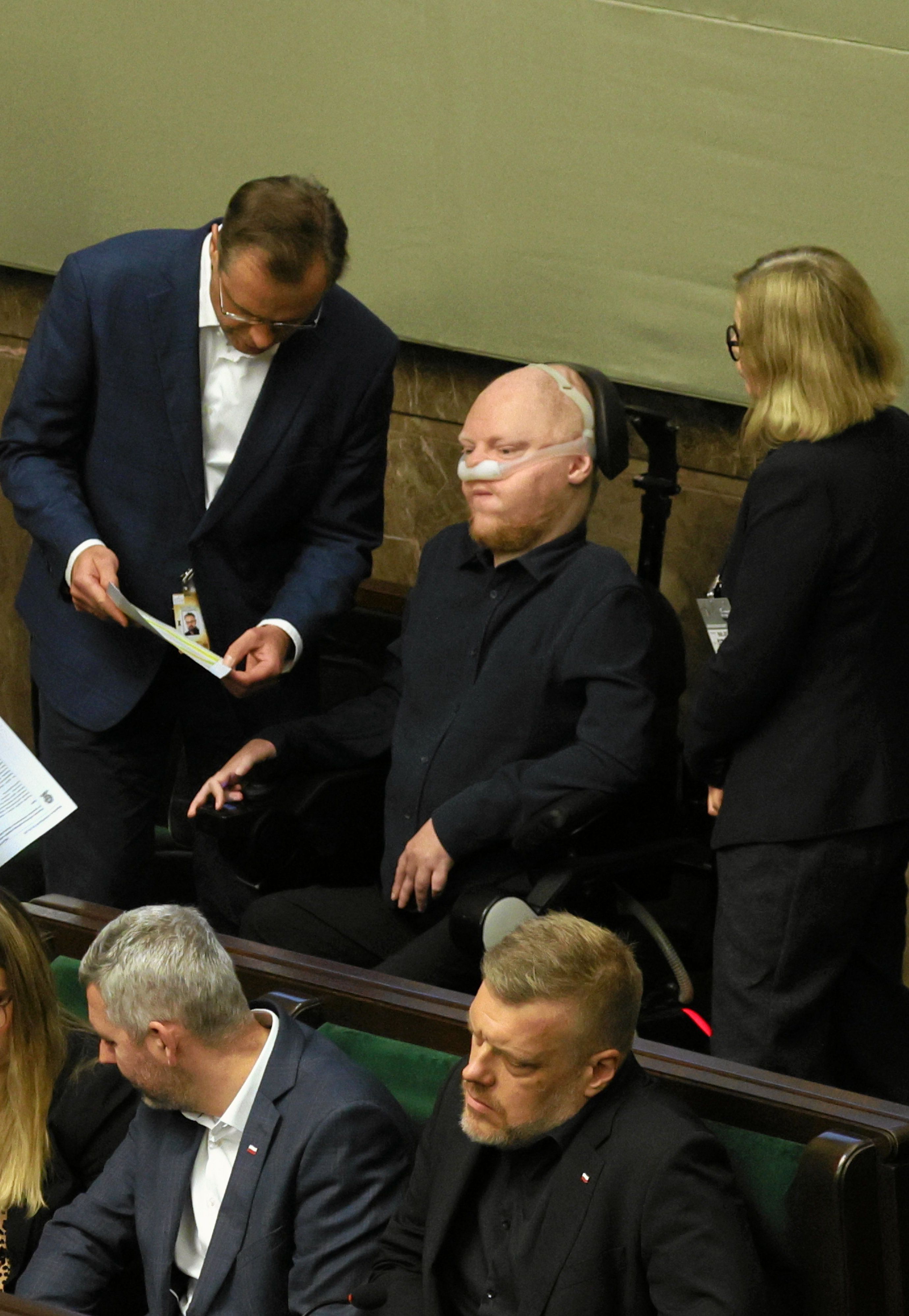 Ludzie w ławach rządowych. Po środku młody mężczyzna na wózku i z aparatem wspomagającym odychanie