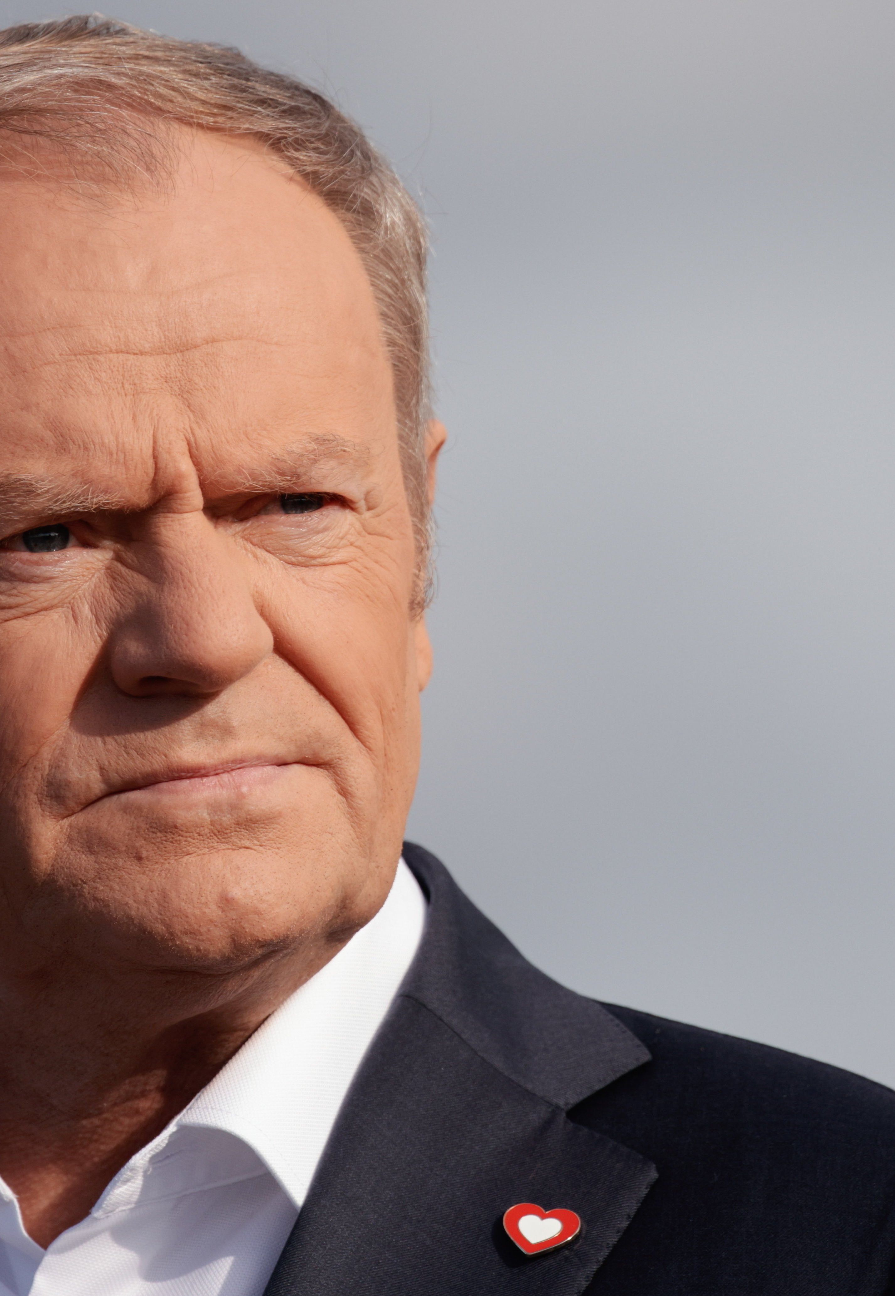Starszy mężczyzna, z krótkimi siwo-blond włosami, w białej koszuli, czarnej marynarce, do której klapy przypięte jest małe, biało-czerwone serduszko, patrzy ze skupieniem w dal.