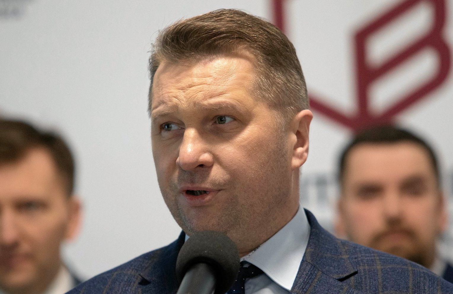 Minister edukacji i nauki Przemysław Czarnek przemawia na konferencji prasowej do mikrofonu. W tle dwóch innych mężczyzn, ich twarze są nieostre.