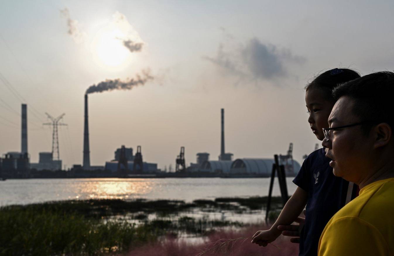 Mężczyzna, Chińczyk, trzyma dziecko na rękach, stoi nad brzegiem rzeki, w oddali kominy elektrowni węglowej