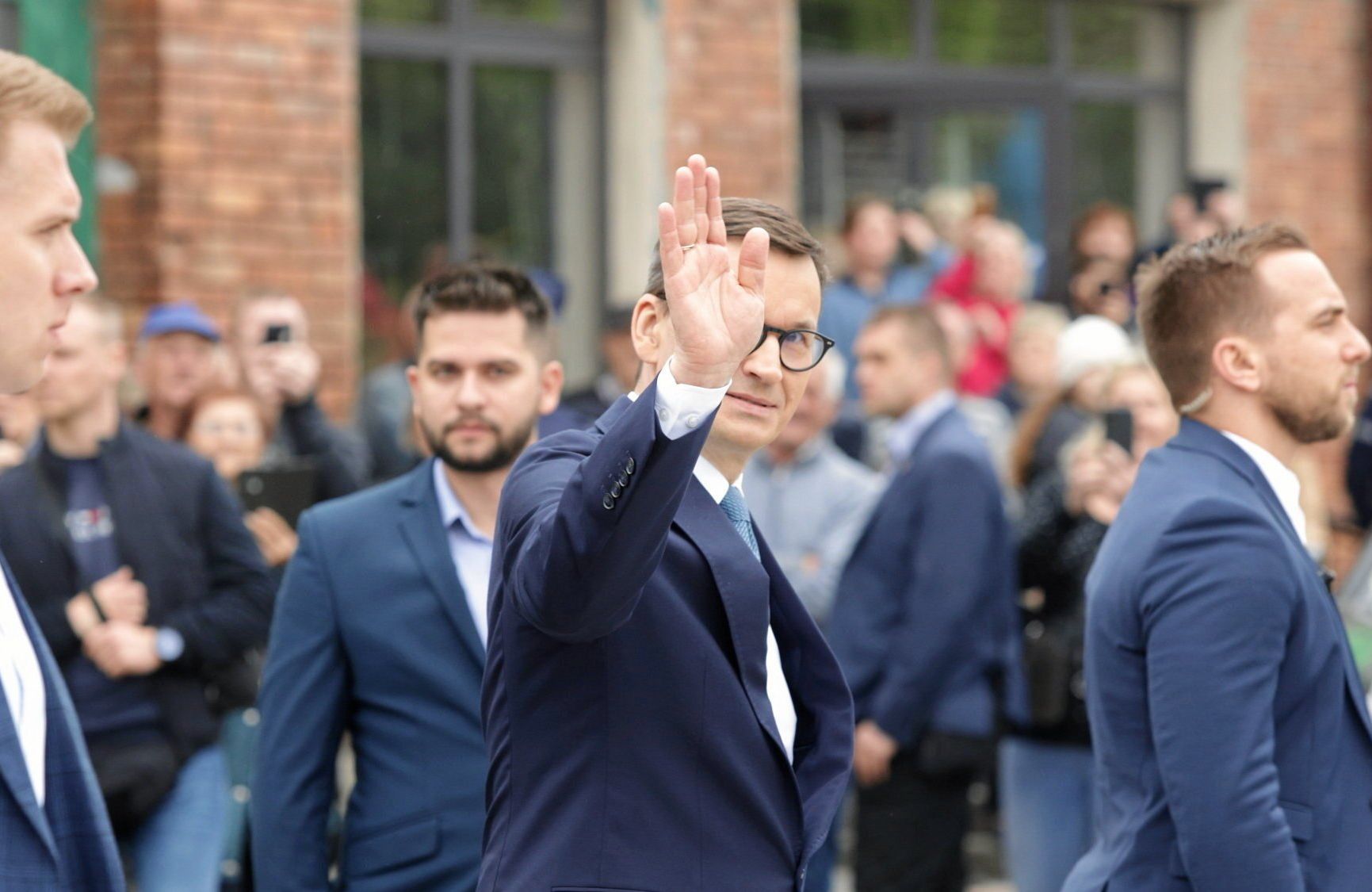 Mężczyzna w garniturze idzie przez plac w otoczeniu innych mężczyzn, macha w kierunku obiektywu