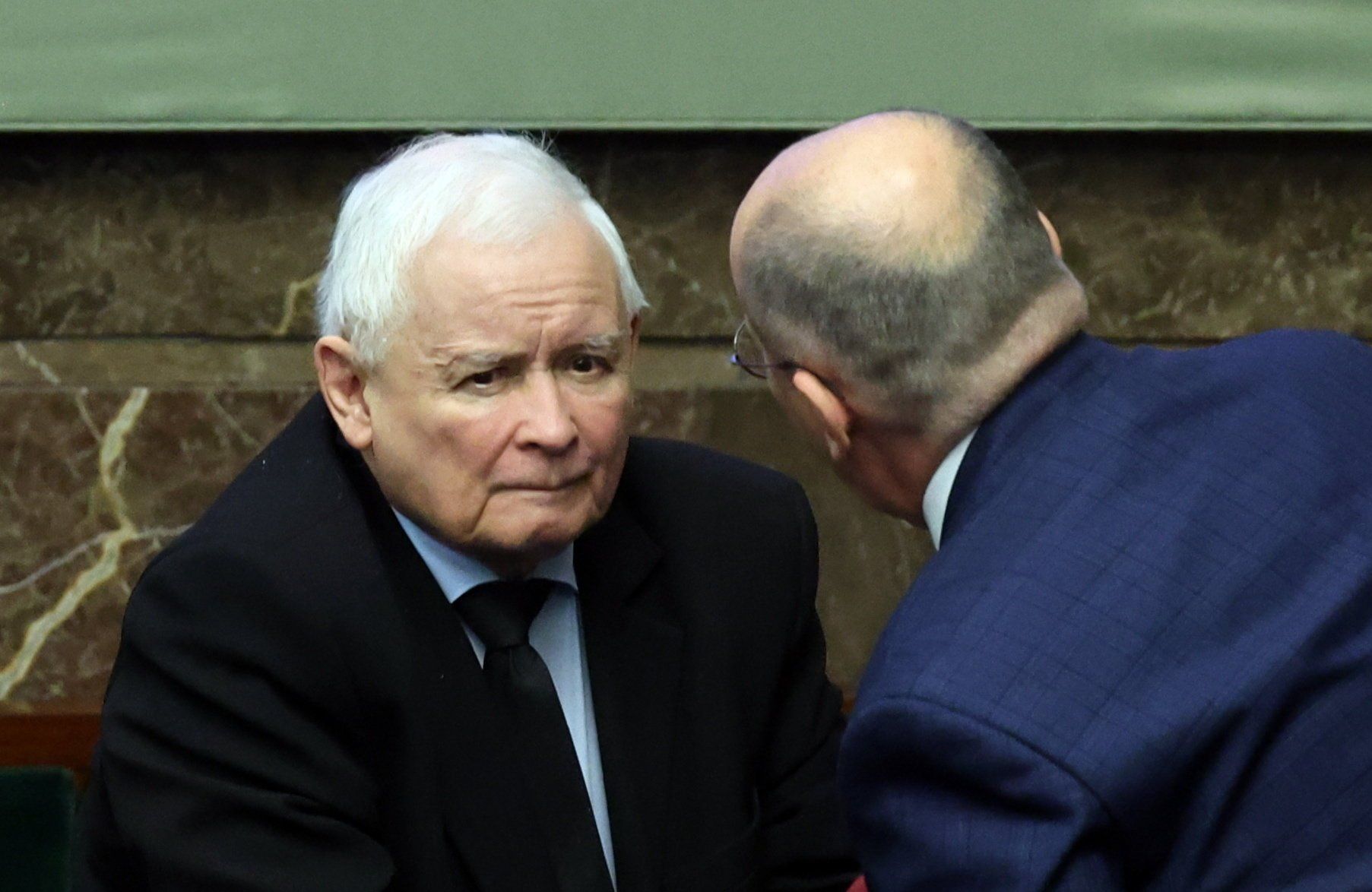 Prezes PiS Jarosław Kaczyński siedzie w ławach rządowych w Sejmie. Pochyla się nad nim jakiś mężczyzna