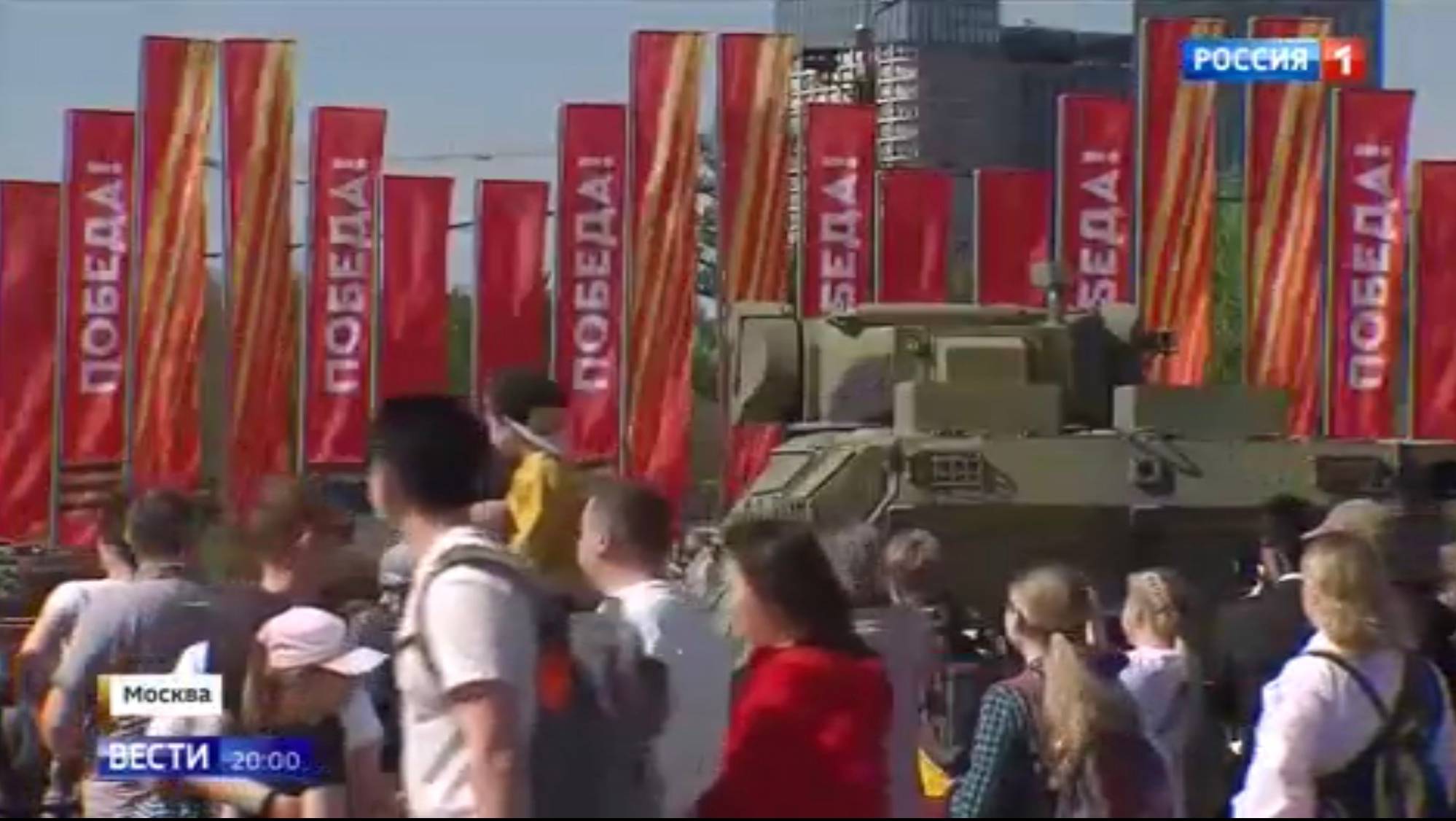 Ludzie oglądają sprzęt wojskowy, w tle - czerwone flagi z rosyjskim napisem 'zwycięstwo"