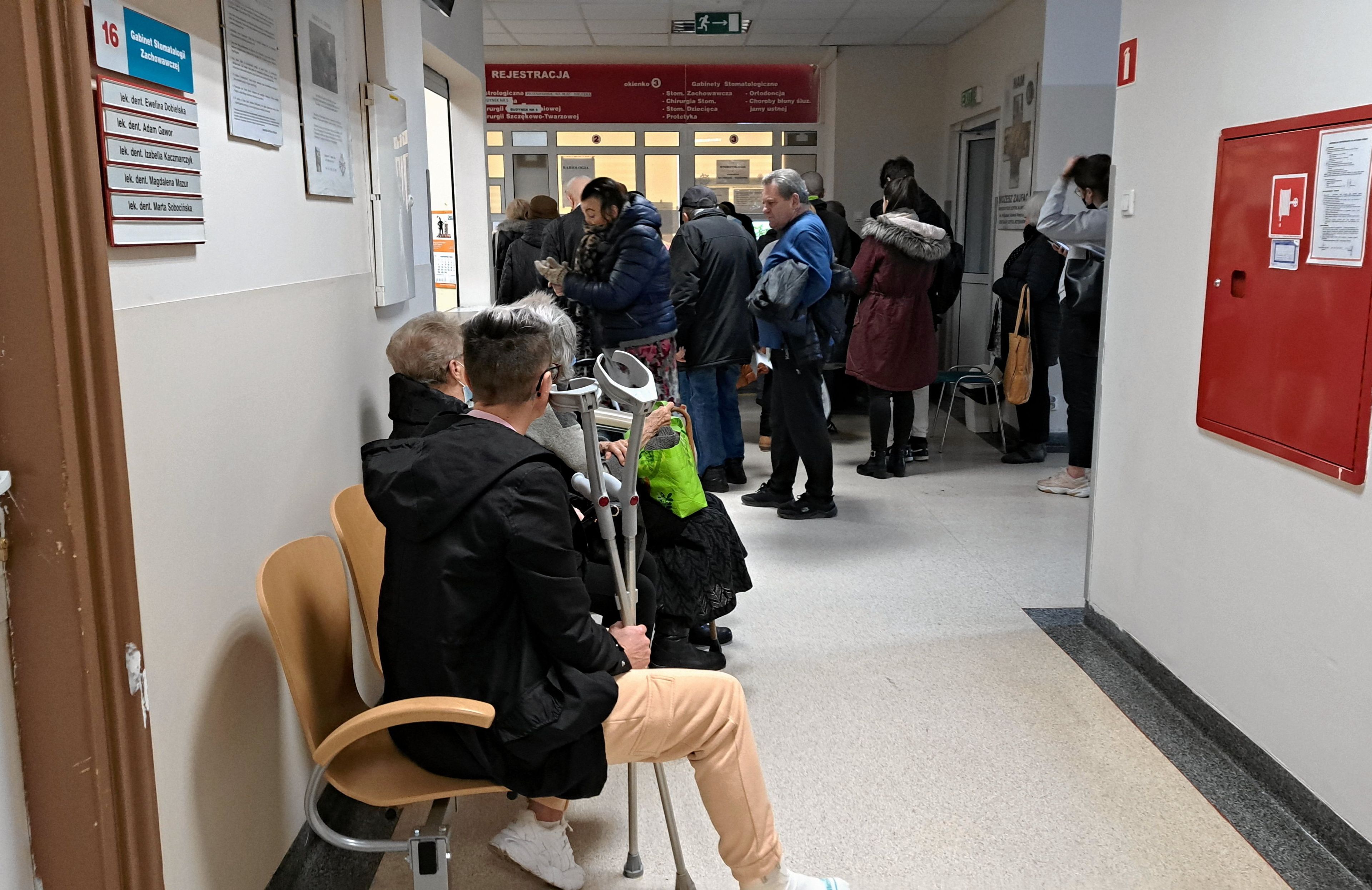 Korytarz przychodni, na pierwszym plane siedzi na krześle mężczyzna i trzyma w ręku kule, w głębi korytarza kilkanaście osób stoi w kolejce do okienek rejestracji