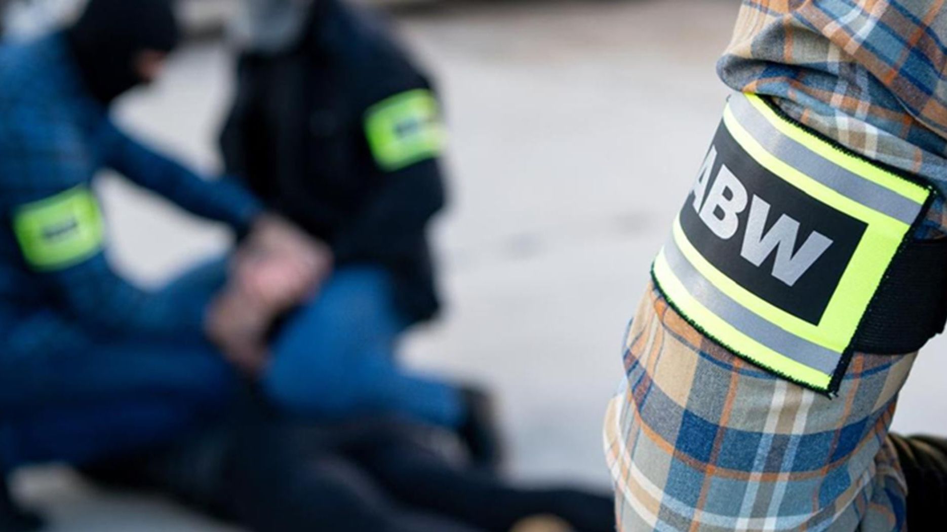 Na pierwszym planie ręka mężczyzny, w koszuli wkratę i z opaską z napisem ABW. W tle dwóch nieostrych funkcjonariuszy ABW podczas zatrzymania osoby.