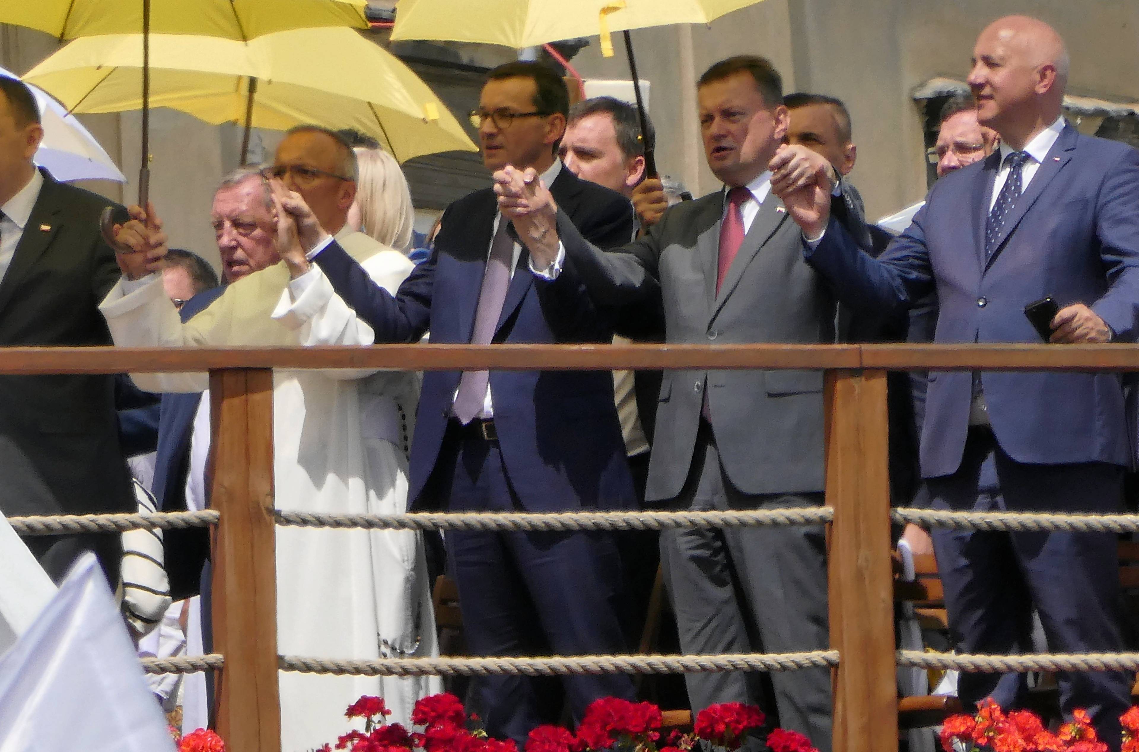 Mateusz Morawiecki, Mariusz Błaszczak i zakonnik w białym habicie trzymają się za ręce, nad nimi żółte parasole