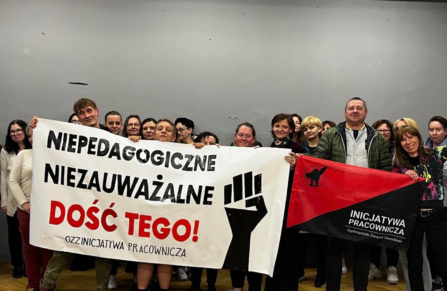 Grupa około 30 osób stoi na parkiecie, na tle szarej ściany, za transparentem z napisem "Niepedagogiczne, niezauważalne. Dość tego!"