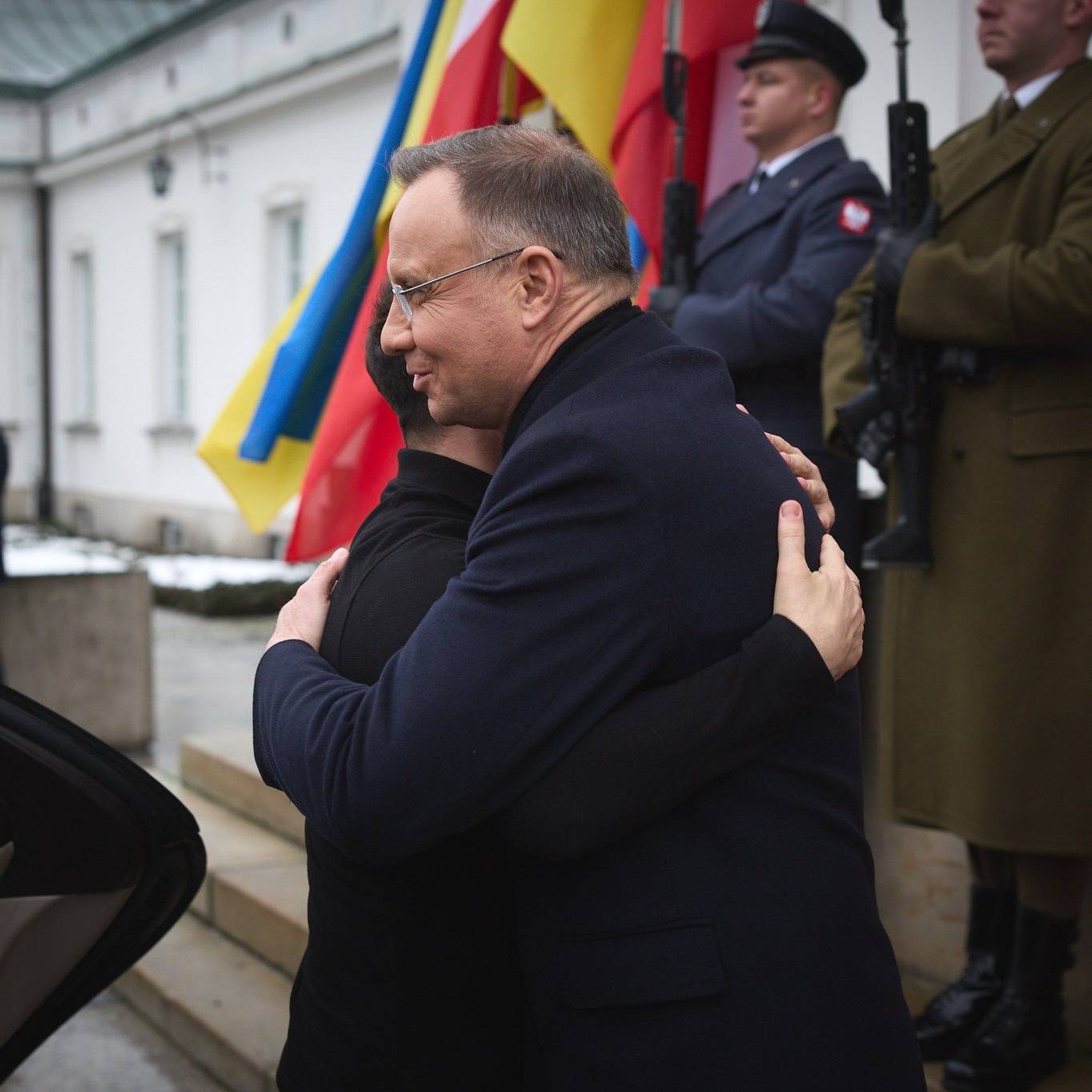 na zdjęciu jest czterech mężczyzn. Mężczyzna w czarnym płaszczu ściska mężczyznę w czarnej bluzie. Z tyłu stoi dwóch żołnierzy w mundurach polskiej armii