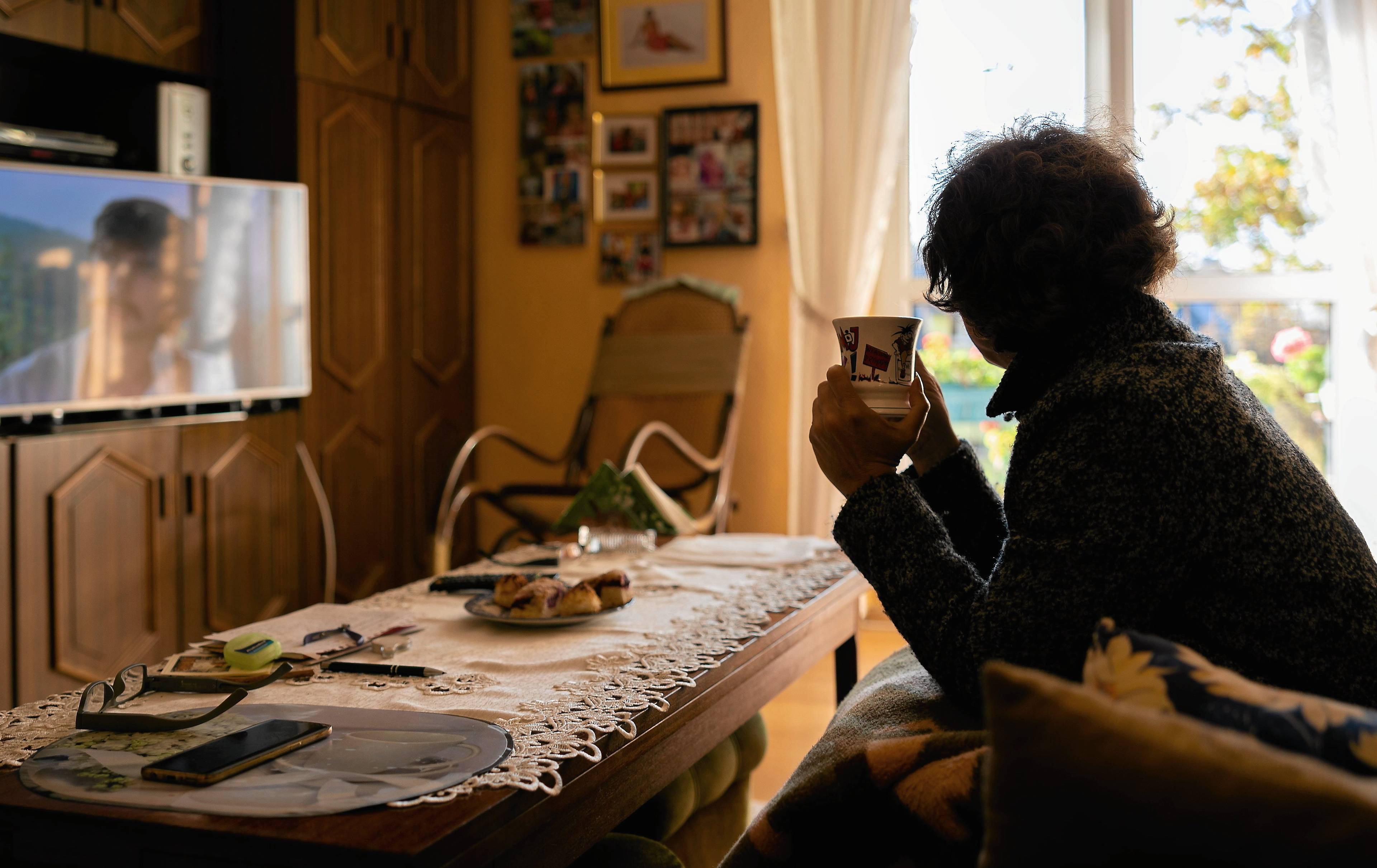 Kobieta siedzi na kanapie w mieszkaniu, jest przykryta kocem w dłoniach trzyma kubek z herbatą