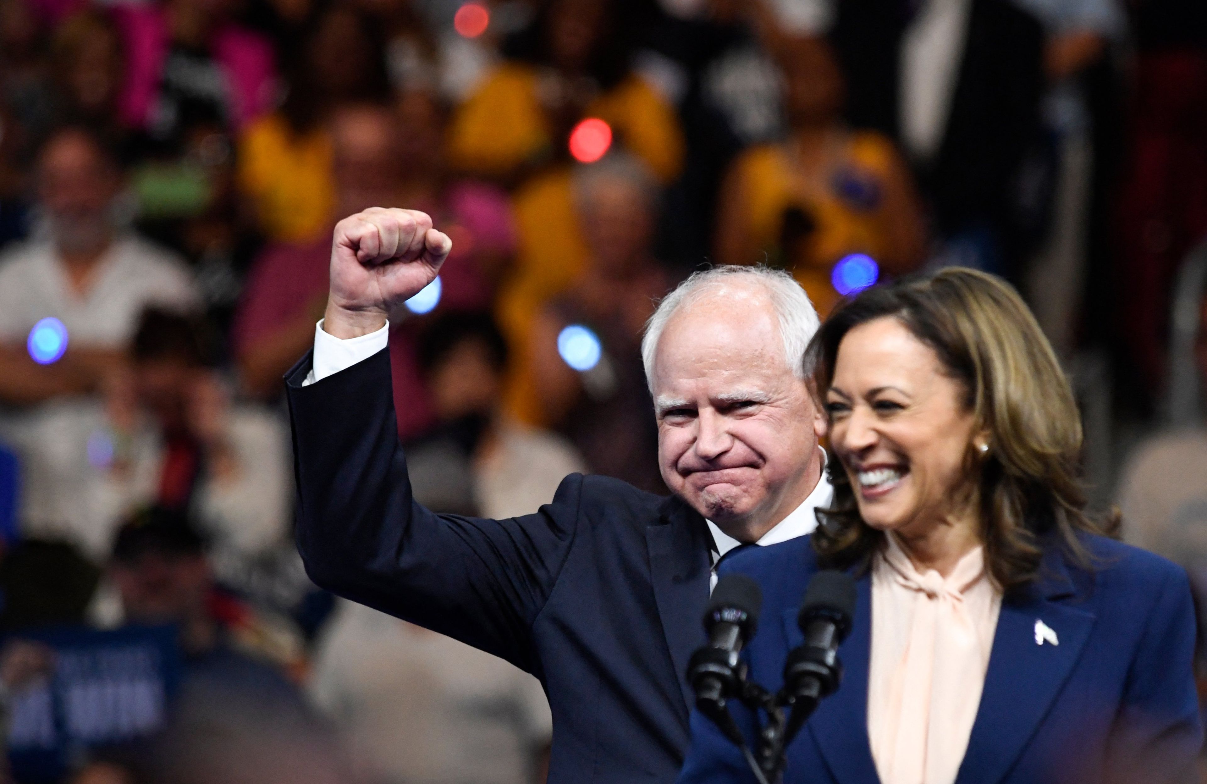 Roześmiana kobieta (Kamala Harris), za nią mężczyzna z podniesioną zwycięsko pięścią (Tim Walz). W tle tłum