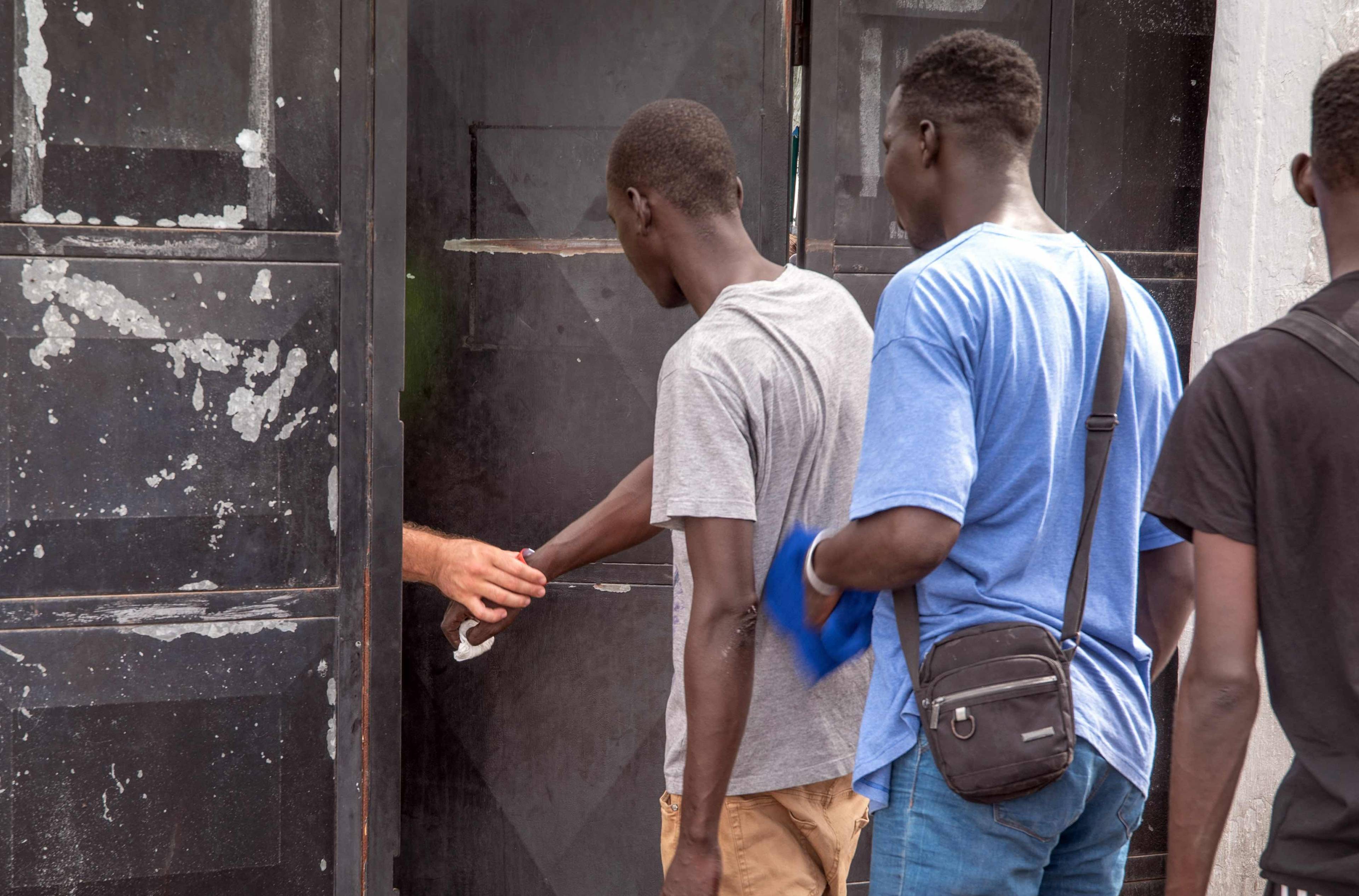 Migranci z Afryki stoją w kolejce przed wejściem do centrum recepcyjnego. W uchylonych drzwiach widać rękę pracownika. Wyspy Kanaryjskie