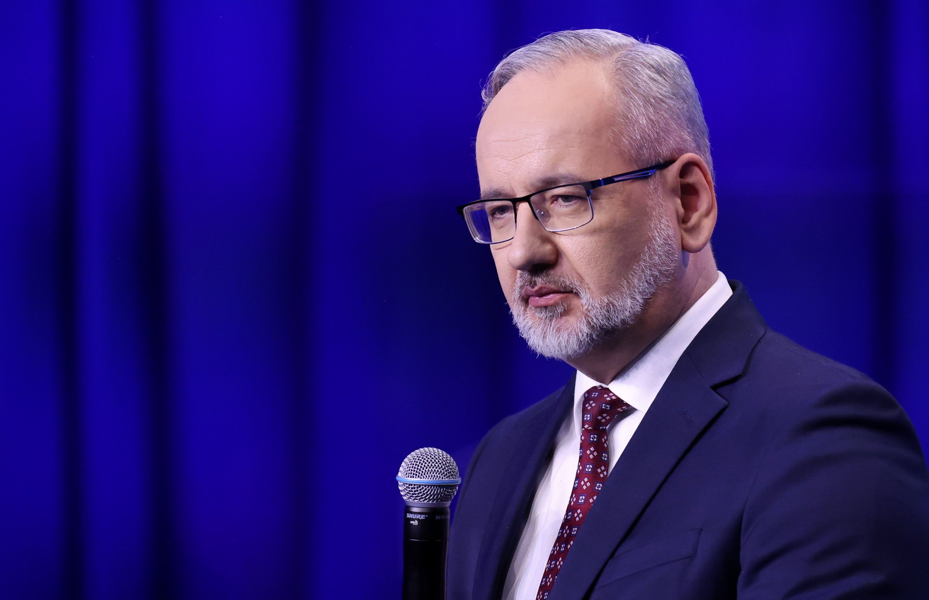 Minister zdrowia stoi w garniturze na niebieskim tle. W ręku trzyma mikrofon