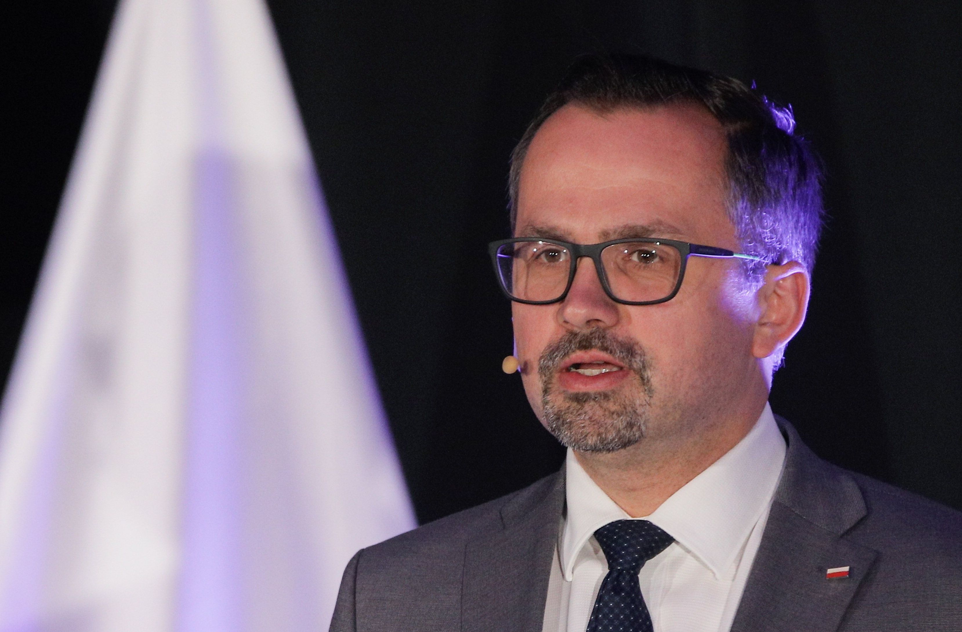 Mężczyzna w jasnej marynarce i okularach, na tle polskiej flagi