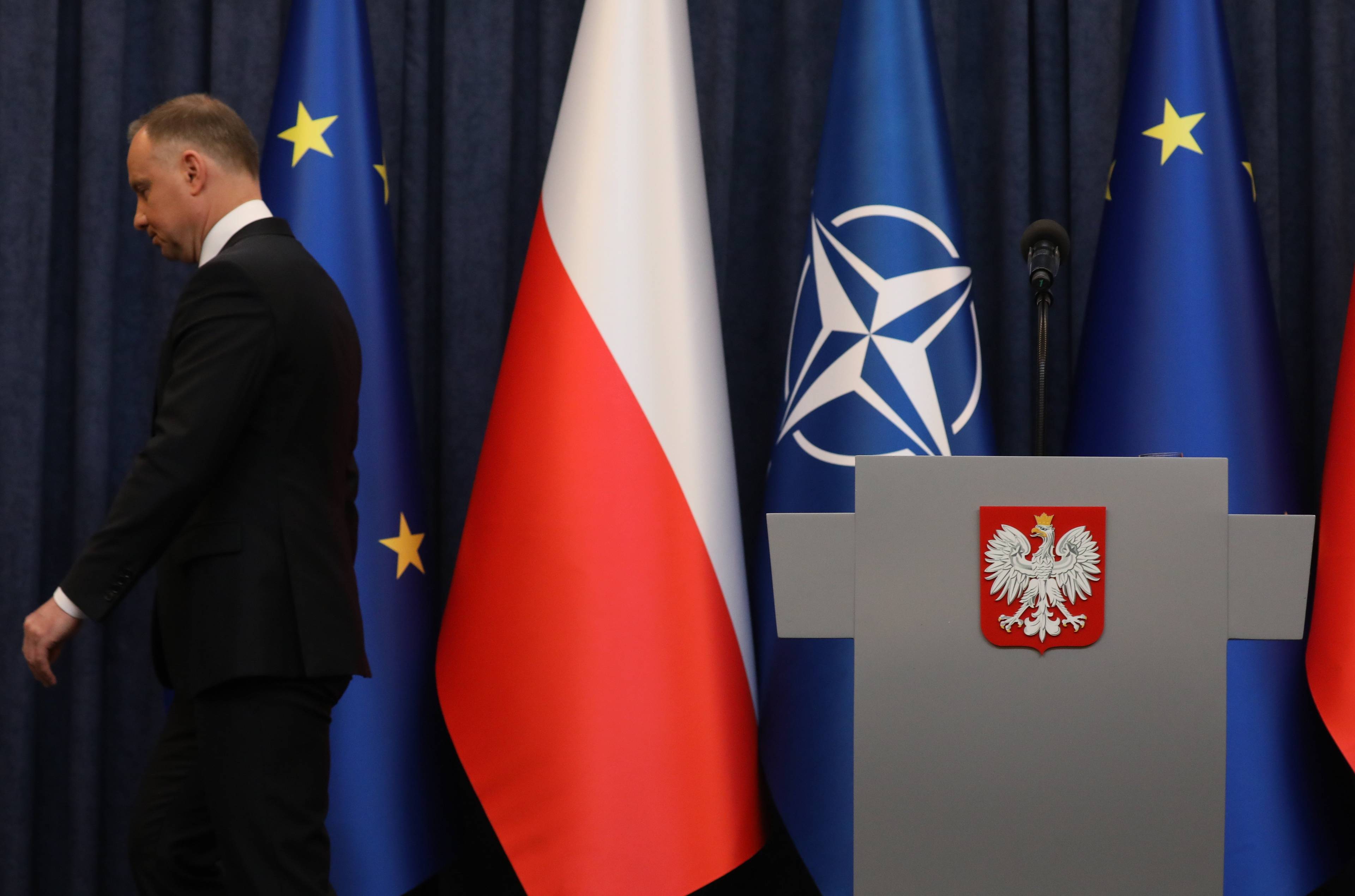 Andrzej Duda schodzi z podestu w sali konferencyjnej w Pałacu Prezydenckim, jest odwrócony plecami do mównicy, w tle flagi Polski, UE i NATO