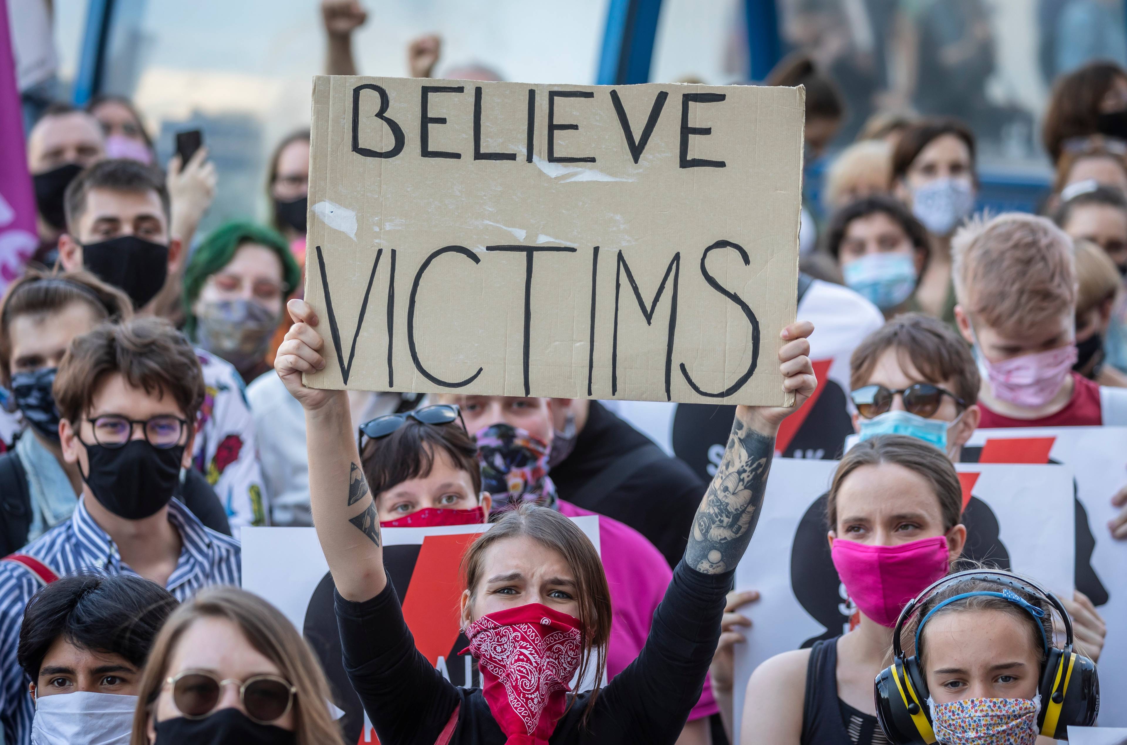 kobieta z czerwoną chustą na twarzy trzyma transparent z napisem na tekturze "Believe victims"